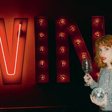 wine in red neon and marquee letters with a woman standing in front. she has red hair and a silver top. she holds a wine glass and mini disco ball.